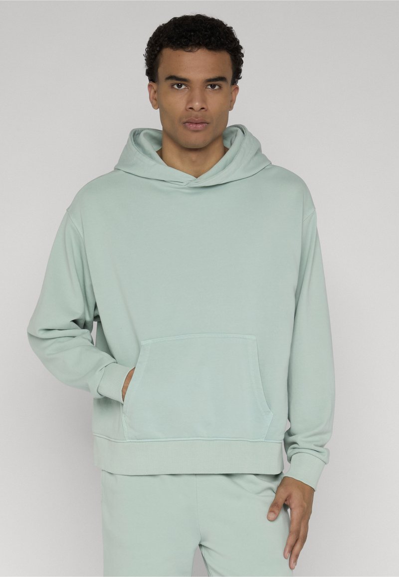 Jeune homme portant un sweat à capuche vert clair avec une poche avant, debout devant un fond gris clair uni.
