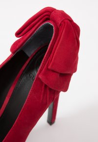 Chaussure à talon haut en suede rouge avec un détail en nœud à l'arrière, doublure intérieure noire et un talon fin et brillant noir.