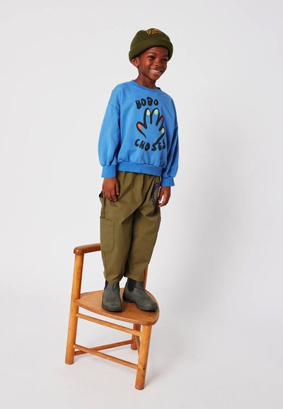 Felpa blu con grafica a mano colorata, pantaloni larghi verde oliva e stivali di camoscio grigi. Bambino in piedi su una sedia di legno.