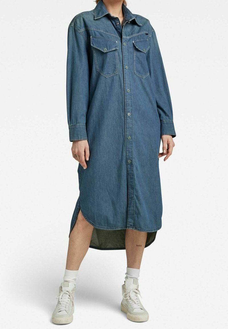 Personne portant une robe chemise en denim bleu à manches longues, boutonnée avec un ourlet fendu, associée à des chaussettes blanches et des baskets blanches montantes.
