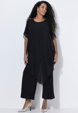 Tunique noire en chiffon au design fluide, à manches courtes et avec des accents aux épaules. Associée à un pantalon noir ample et des talons argentés.