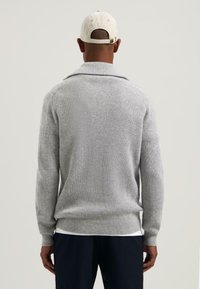 Grijze geribbelde trui met sjaalkraag, lange mouwen en een getailleerde taille. Draagt over een wit shirt, gecombineerd met donkere broeken. Witte pet op het hoofd.