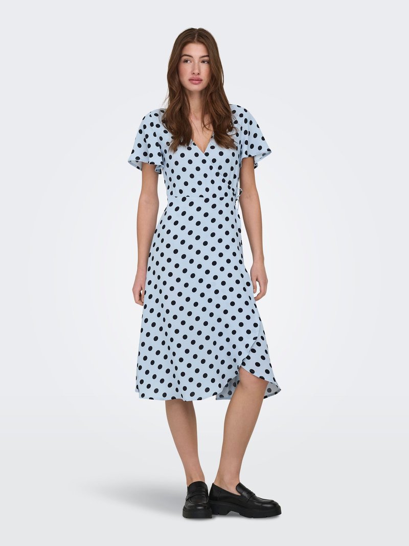 Kvinne står iført en lys blå midikjole med svarte prikker, flagrende ermer, omslagskjørt og svarte loafers mot ensfarget bakgrunn.