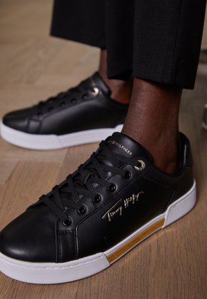 Czarne niskie trampki z białymi podeszwami i złotym logo Tommy Hilfiger, noszone z czarnymi spodniami na drewnianej podłodze.