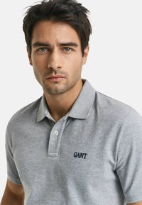 Junger Mann mit dunklen Haaren, der ein hellgraues GANT-Polohemd trägt und mit Blick zur Kamera vor einem schlichten weißen Hintergrund steht.