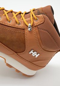 Bota de senderismo de cuero marrón con acentos de malla, cordones amarillos, herrajes plateados y una suela de goma naranja, con detalle del logo en el costado.