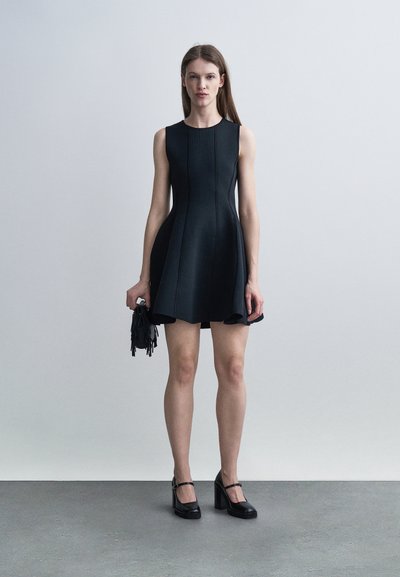 Γυναίκα που φοράει μαύρο αμάνικο φόρεμα και μαύρα τακούνια block, κρατώντας μια μικρή μαύρη τσάντα με φούντες, στέκεται μπροστά σε έναν απλό ανοιχτόχρωμο φόντο.