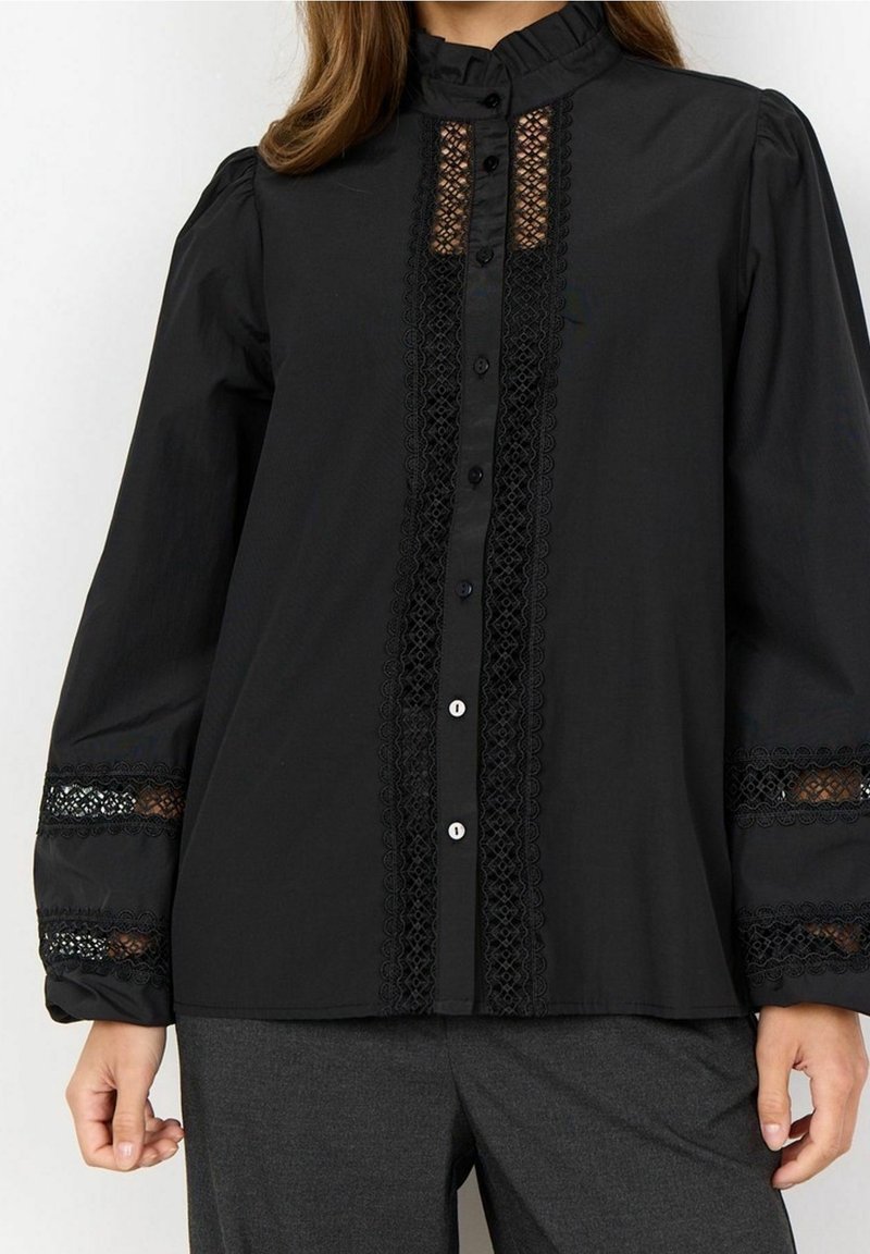 Femme portant un chemisier noir à manches longues avec un bord en dentelle sur le devant et les manches, boutonné devant, associé à un pantalon gris foncé.