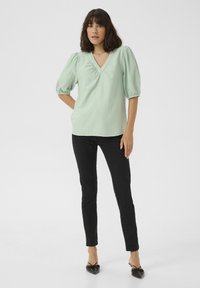 Blouse vert clair texturée avec un décolleté en V et des manches bouffantes, associée à des pantalons noirs ajustés et des chaussures noires à bout pointu.