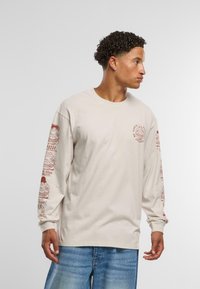 Beige langermet bomull T-skjorte med røde grafiske design på begge ermene og en liten logo på fronten. Avslappet passform.