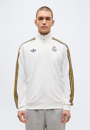 Mladi moški, ki nosi belo športno jakno z zadrgo, z modrimi in rumenimi črtami na rokavih ter s logotipoma Real Madrida in Adidasa, stoji pred belim ozadjem.