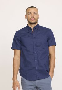 Chemise courte à manches en navy avec un col à boutons, des accents de boutons clairs et un logo subtil sur la poitrine ; tissu texturé avec une finition lisse.