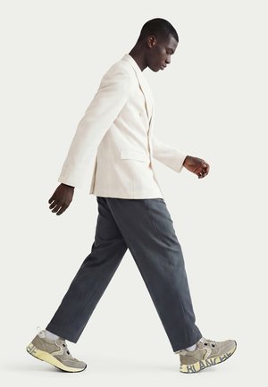 Man walking wearing a cream blazer, dark gray cropped pants, and gray-beige sneakers with thick sole and text on the side.