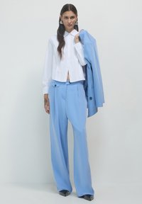 Chemise blanche à boutons raccourcie avec manches longues ; pantalon large bleu clair taille haute ; blazer bleu assorti drapé sur l'épaule.