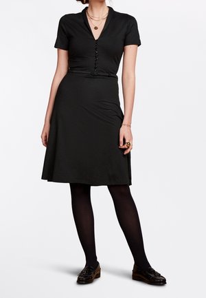 Frau in einem knieumspielenden schwarzen Kleid mit kurzen Ärmeln, schwarzen Strumpfhosen, braunen Slippern und geschichteten goldenen Halsketten, stehend vor weißem Hintergrund.