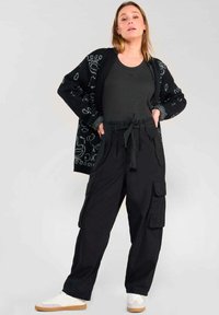 Zwarte cargo broeken met meerdere zijzakken, gecombineerd met een donkere tanktop en een gestreepte zwarte cardigan. Sportieve witte sneakers maken de look compleet.