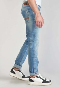 Verbleekte blauwe denim jeans met een slim fit, contrasterende stiksels en een leren label bij de taille; gedragen met wit en zwarte sneakers.