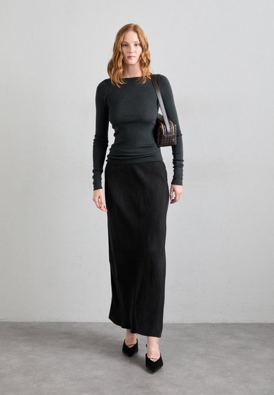 Dark green long-sleeve fitted top paired with a black floor-length skirt. Black heeled shoes with pointed toes and a small textured handbag.