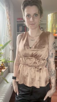Satinový béžový peplum top s výstřihem do V a nařaseným pasem. Hladká textura, bezrukávový design, kombinovaný s černými kalhotami.