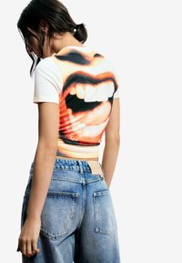 Žena s tmavými vlasmi v pekáči nosí biele krátke tričko s veľkou grafikou otvorených úst na chrbte a voľné modré rifle.