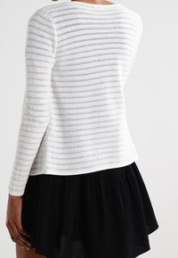 Maglione bianco lavorato a maglia con righe orizzontali grigie, scollatura rotonda e maniche lunghe, abbinato a una gonna nera a strati.