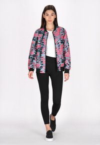 Multicolor bomberjack met een roze, blauwe en zwarte print, gecombineerd met zwarte legging en zwarte sneakers. Glad materiaal en geribbelde manchetten.