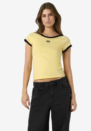 Ung kvinne iført en gul crop-top med korte ermer og svarte kanter, samt høytlivs svarte bukser, står foran en ensfarget bakgrunn.