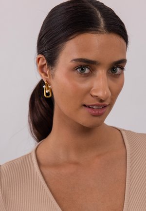 Boucles d'oreilles en or en forme de cercle avec un design superposé, présentant des accents en métal lisse et en perles, portées par une personne aux cheveux sombres sur un fond neutre.