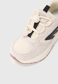 Lys beige sneakers med mesh overdel, suedebeslag, sorte snørebånd og en rund justeringslås. Hvid mellemsål og teksturert gummisåle.