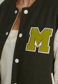 Mørkegrøn varsity-jakke med kviltede hvide ærmer, der har et tekstureret grønt "M"-patch og hvide trykknapper foran.