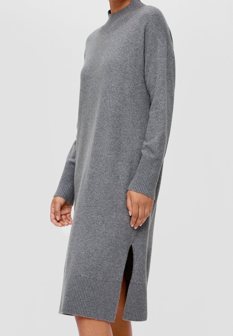 Femme portant une longue robe pull grise en tricot avec des poignets côtelés, un col montant et une fente latérale, debout devant un fond clair uni.