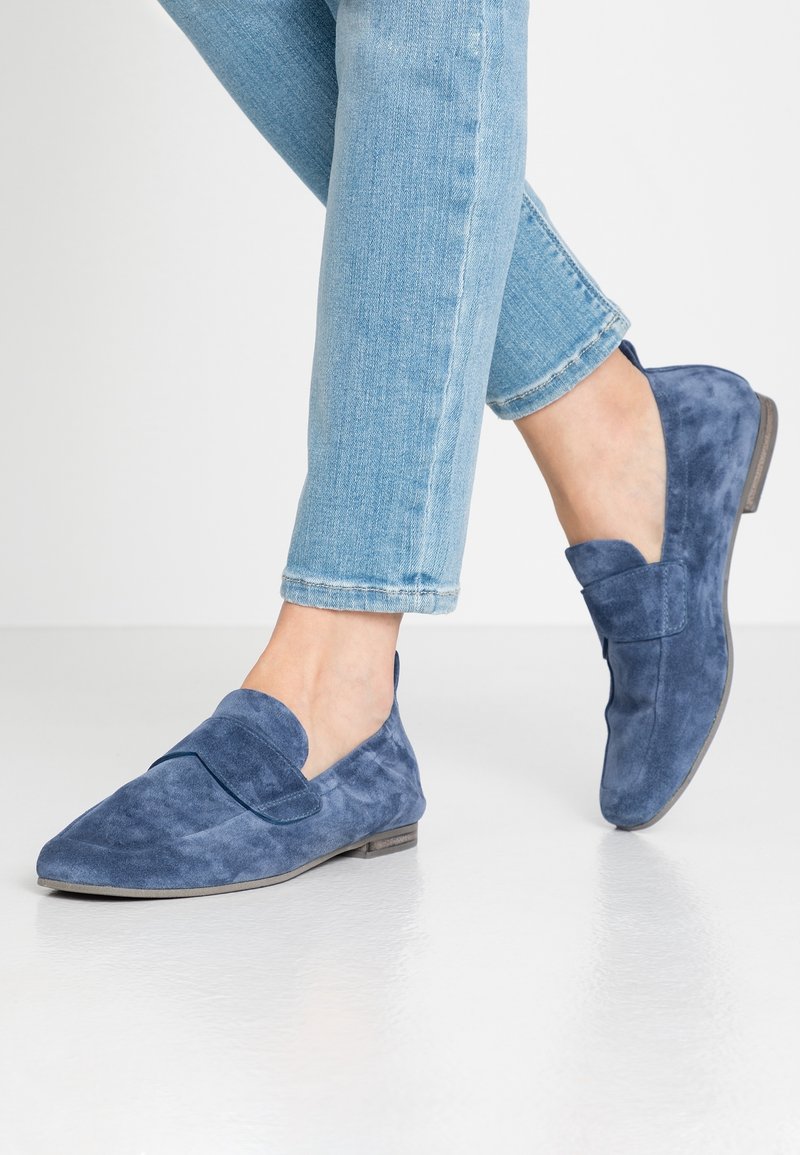 Personne portant un jean bleu clair et des mocassins en daim bleu debout sur un sol blanc réfléchissant.