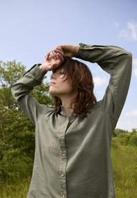Femme aux yeux fermés, étirant les bras au-dessus de la tête, portant une chemise verte boutonnée, debout en plein air avec des arbres et un ciel bleu en arrière-plan.