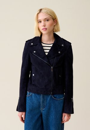 Giacca da moto in suede blu navy con accessori in metallo argentato, tasche con zip e un design del colletto classico. Abbinata a una maglietta a righe e denim.