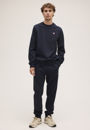 Joven con sudadera azul marino y pantalones deportivos a juego con zapatillas blancas y beige, de pie frente a un fondo claro liso.