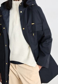 Námořnická parka s kapucí, zlatým kován ím a zipovým uzávěrem, v kombinaci s krémovým pleteným svetr. Obsahuje boční kapsy a má uvolněný střih.