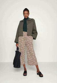 Mulher vestindo uma jaqueta cropped oliva, um suéter de gola alta preto, calças largas florais, chinelos pretos e segurando uma mala tote preta.