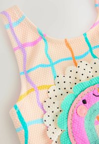 Top de baño colorido con una superficie texturizada de color rosa pálido y un patrón cuadriculado multicolor. Cuenta con un aplique decorativo de dona bordada con lunares.