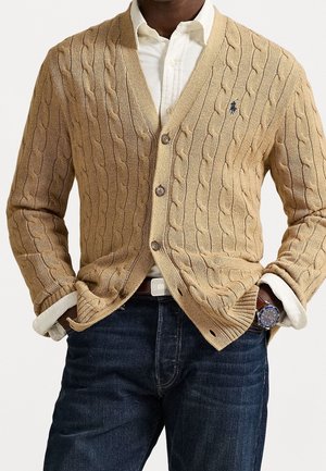Homme portant un cardigan beige à torsades par-dessus une chemise blanche boutonnée, jean bleu, et une montre-bracelet, les mains dans les poches, torse visible.