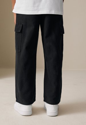 Personne portant un pantalon cargo noir à jambes larges et des baskets blanches, debout sur un sol clair sur fond neutre.