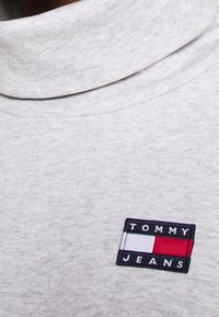 Lichtgrijze coltrui van zacht materiaal, met een gestikte rechthoekige logo waarop "TOMMY JEANS" staat en een blauw-rood-witte kleurenschema.