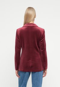 Burgundské sametové sako s klopovým límcem, zapínáním na knoflíky a rozparkem vzadu. Nositelný přes světle modré džíny, které ukazují hladkou texturu.