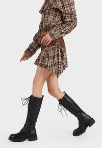 Stivali militari neri alti fino al ginocchio con allacciatura frontale e suola texturizzata. Materiale in pelle liscia, design robusto con zip laterale e supporto per la caviglia.