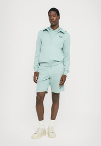 Lys myntgrønn zip-up hettegenser med spiss krage og matchende shorts. Har logodetaljer, myk stoff og hvite joggesko med polstrede såler.