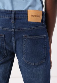 Denim jeans in dark blue with a leather patch labeled "ONLY & SONS" on the waistband, featuring standard back pockets and contrast stitching.