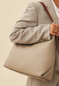 Beige leather handbag with a smooth texture, gold hardware accents, and a brown shoulder strap, featured against a neutral background.