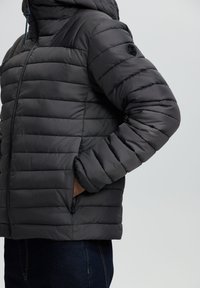 Mand iført en mørkegrå quilted jakke med hætte, hånd i lommen, parret med mørkeblå jeans foran en ensfarvet baggrund.