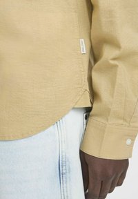 Beige košile s dlouhým rukávem z texturované látky. Má zakulacený lem a manžety na knoflíky. Nápis viditelný na spodním švu. Nosí se s lehkými džíny.
