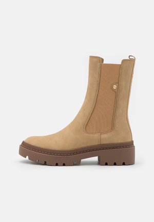 Tan Chelsea boots made of suede with elastic side panels, a rounded toe, and a chunky brown rubber sole featuring textured accents.