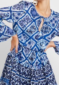 Modré šaty s potiskem obsahující řasený pas, dlouhé rukávy a výstřih ve tvaru klíčové dírky. Design zahrnuje geometrické tvary v různých odstínech modré.